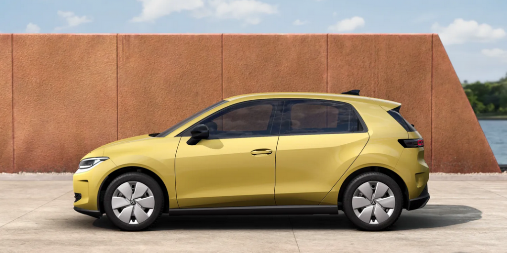 Side view of a yellow compact hatchback car parked on a concrete surface with a brown wall behind it.