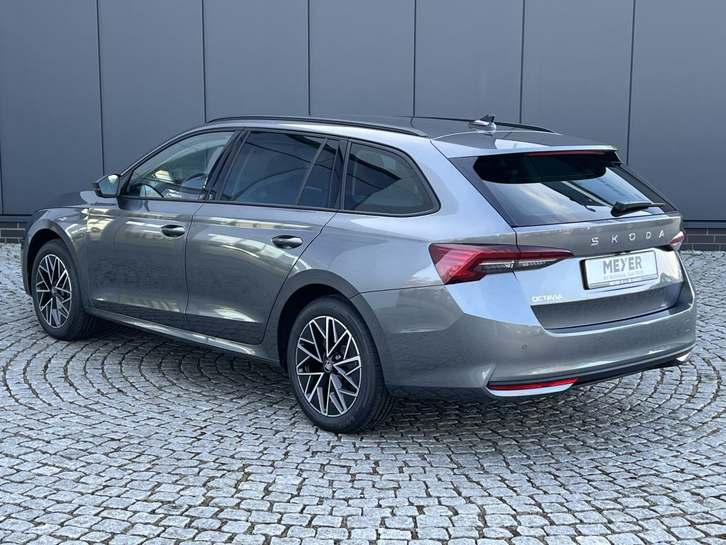 Side-rear view of a silver Skoda Octavia station wagon parked on a cobblestone driveway in front of a gray wall.