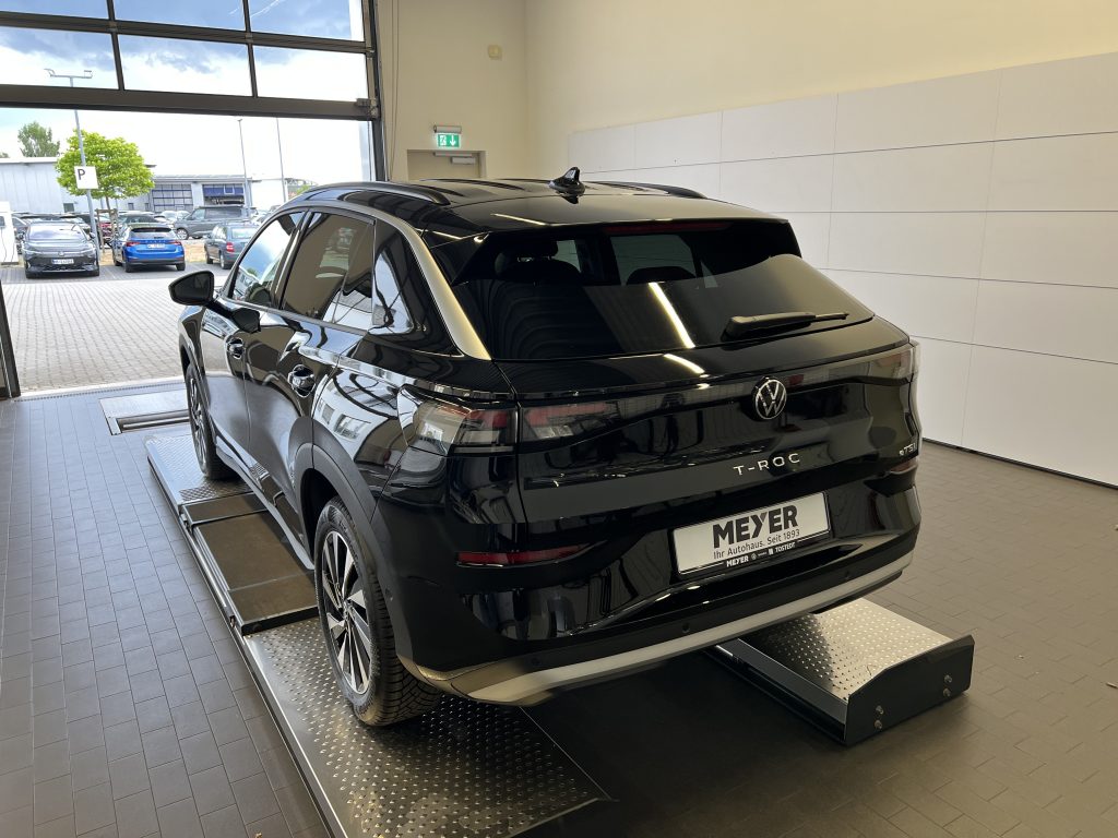 Black Volkswagen T-Roc SUV on a raised platform inside a car showroom, viewed from the rear‑quarter with reflections on its glossy surface. Outside, other vehicles are visible through large showroom doors.
