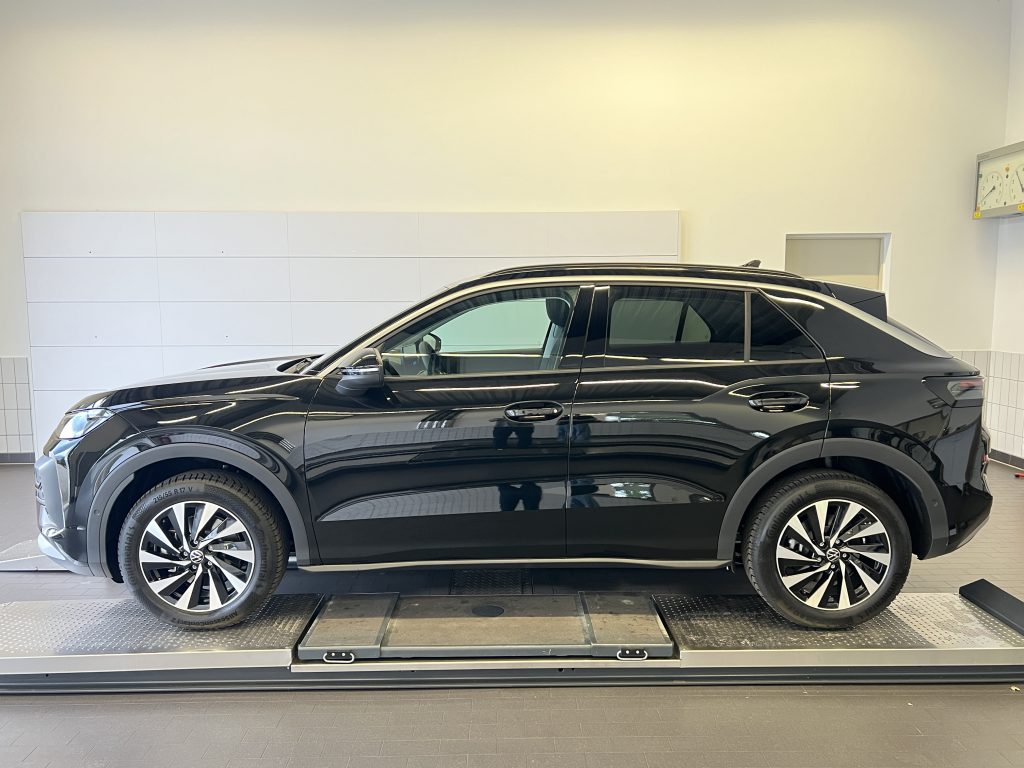 Black SUV in a showroom, side view mounted on a platform with white tiled wall behind and a clock on the right wall.
