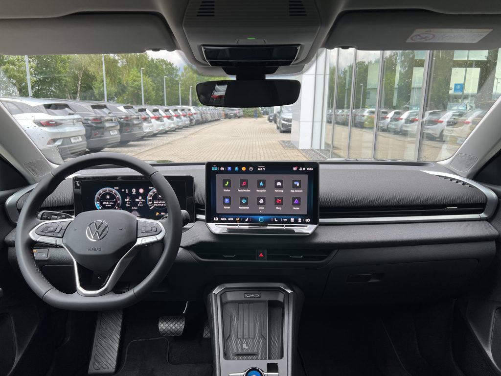 Interior view of a car cabin with a large central touchscreen and digital instrument cluster from the driver's seat, parked at a lot.