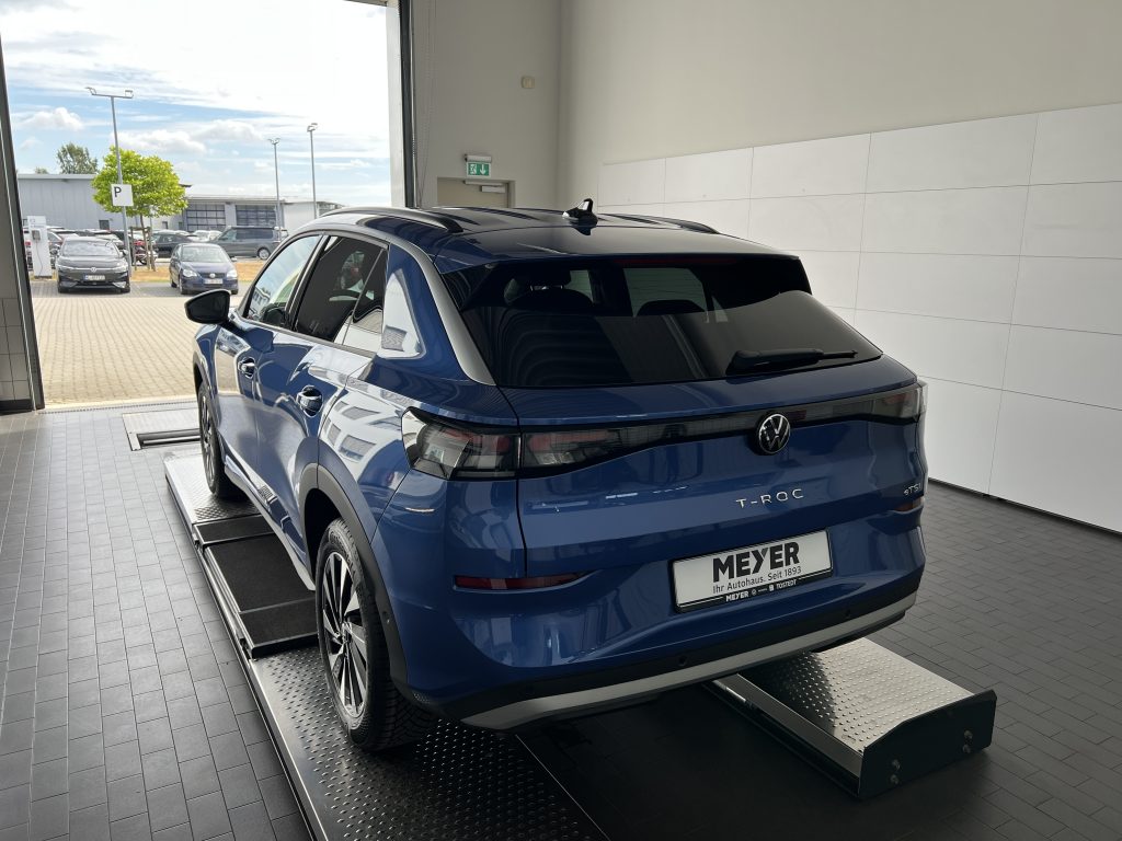 Blue Volkswagen T-Roc SUV on a raised platform inside a showroom, with large glass doors and parking lot visible outside