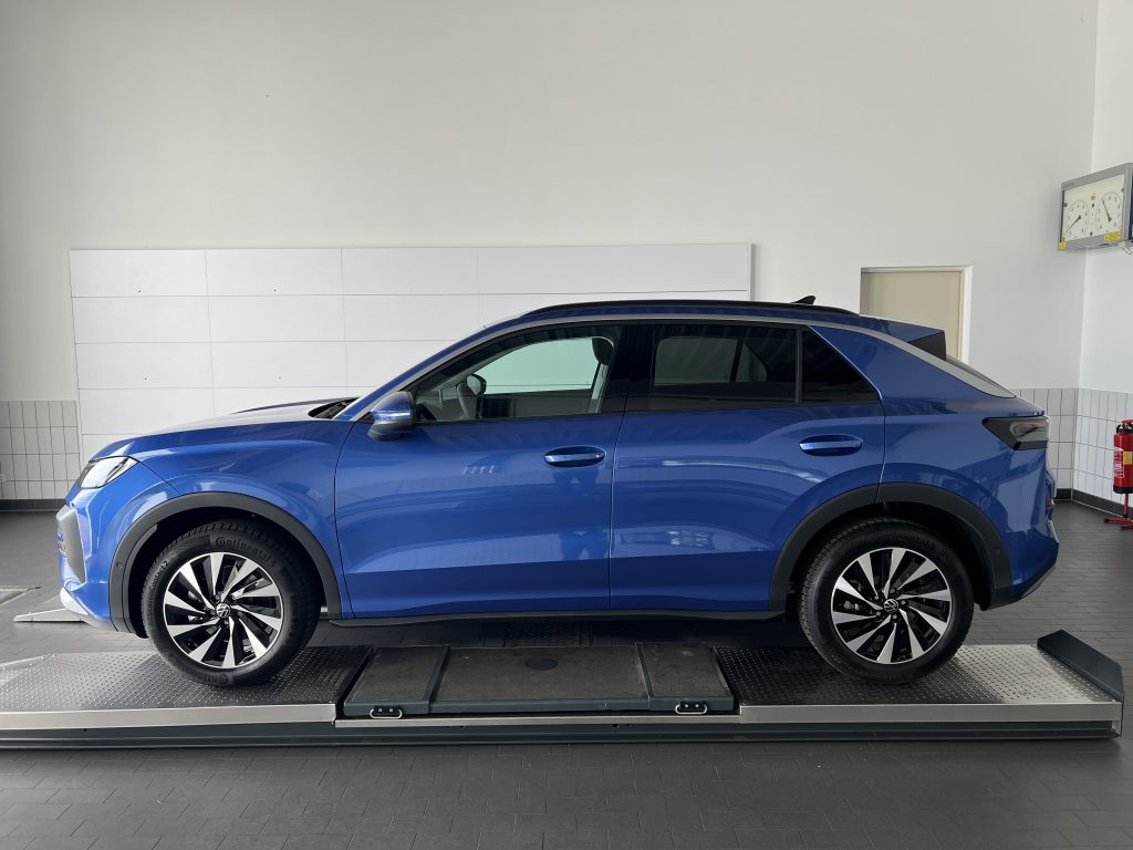 Blue Volkswagen SUV in a showroom, partially on a lift, with white tiled walls and a clock on the right corner visible.