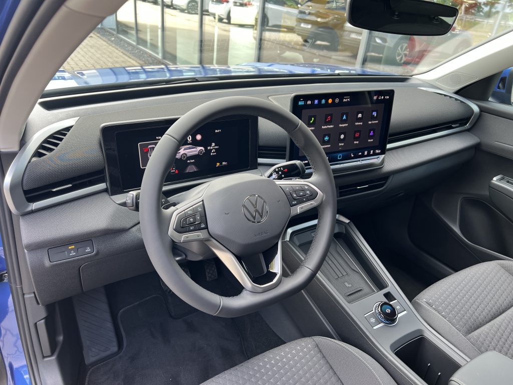 Interior of a car cockpit with a digital instrument cluster to the left and a large central touchscreen on the dashboard, steering wheel in the foreground.
