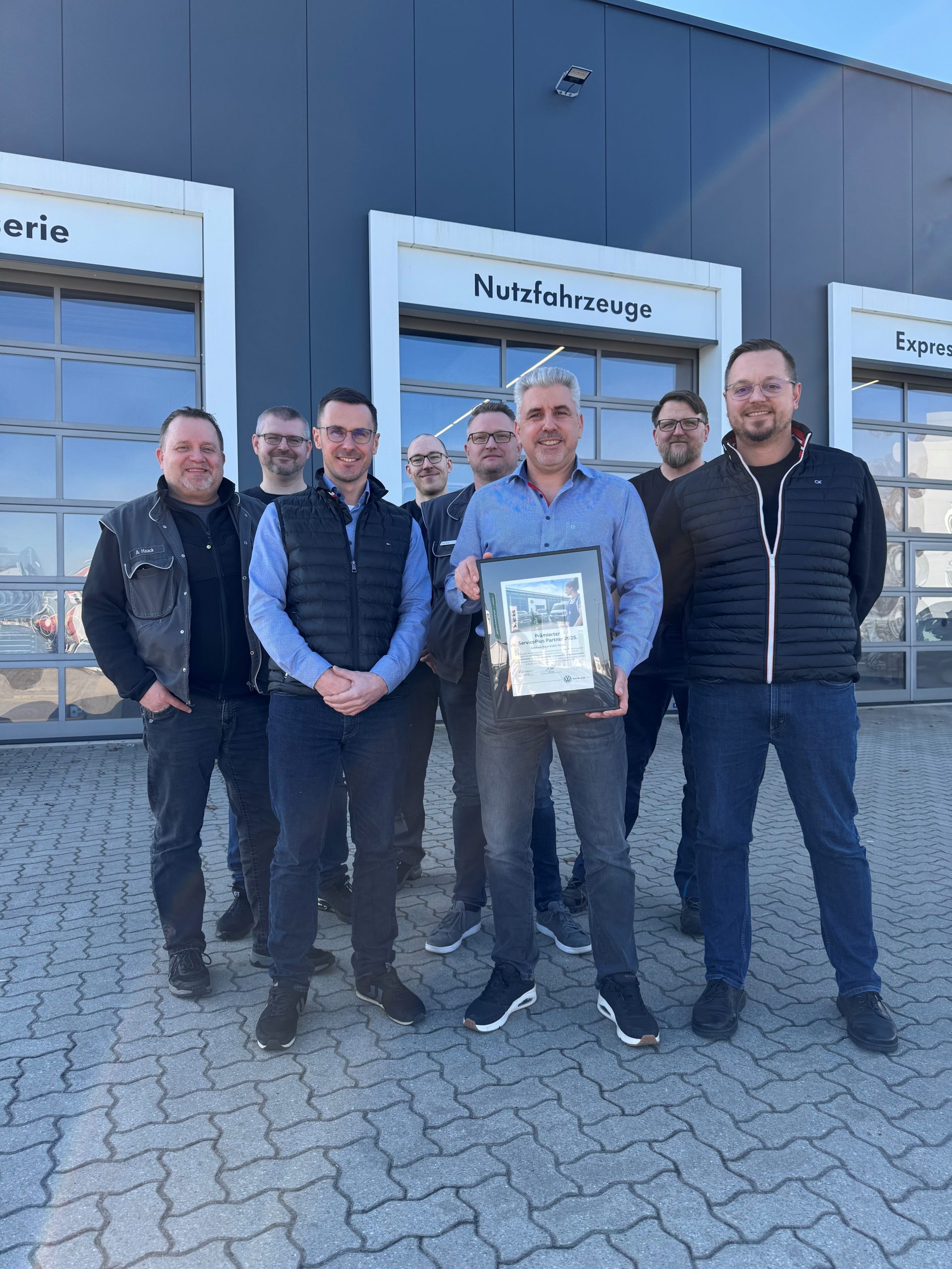 Eight men posing outside a modern automotive service center; the front-and-center man holds a framed certificate or award while others stand behind him in casual workwear.
