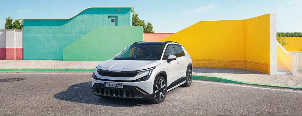 White compact SUV parked on a sunny street against bright, geometric teal, green, and yellow walls in an urban setting.”,
