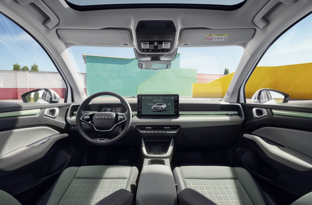 Front-seat view of a Skoda car interior with steering wheel, large central touchscreen, overhead console, and sunroof; colorful exterior mural visible through windows.