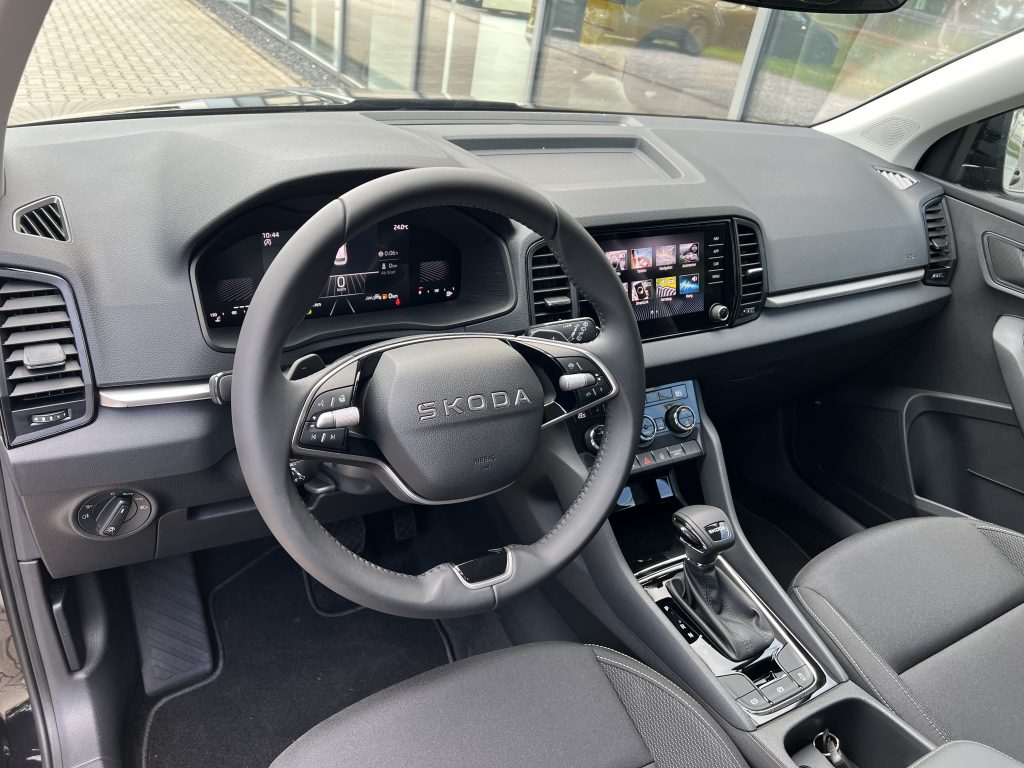 Steering wheel with Skoda badge in a modern car interior, digital instrument cluster behind it and a touchscreen infotainment system to the right.