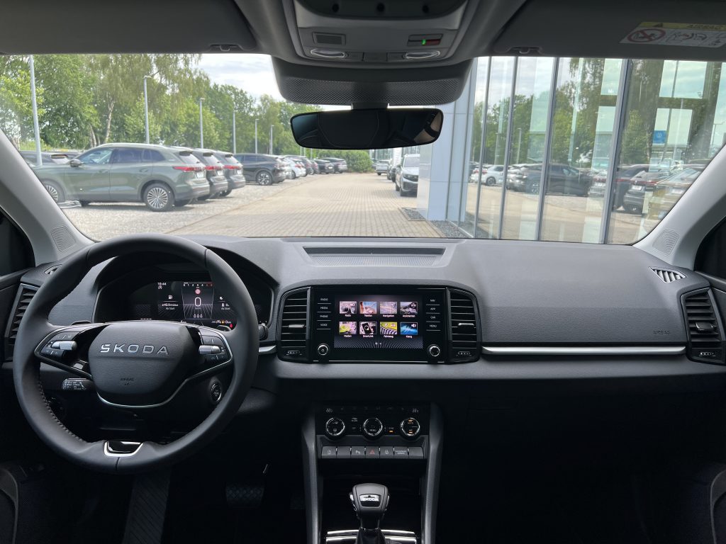 Inside view of a Skoda car cockpit with steering wheel, digital instrument cluster, center touchscreen, gear lever, and rearview mirror; showroom outside through windows.