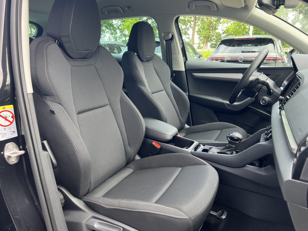 Front interior of a car showing two black fabric seats, center console, and steering wheel with dashboard; parked cars visible outside the windows.