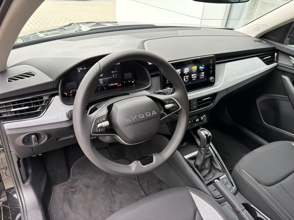 Interior of a modern Skoda car from the driver's side, showing a digital instrument cluster, a large central touchscreen, and a leather-wrapped steering wheel with controls
