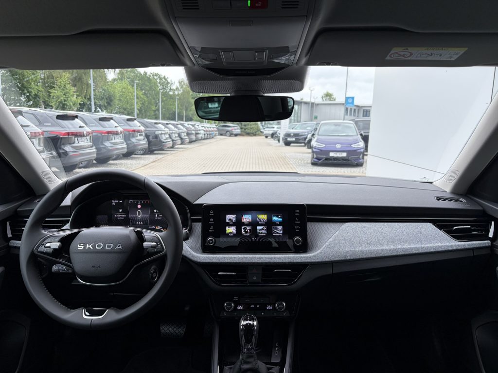 View from the driver’s seat of a Skoda: steering wheel, large central touchscreen, gear shifter, and rearview mirror, with cars parked outside through the windshield.