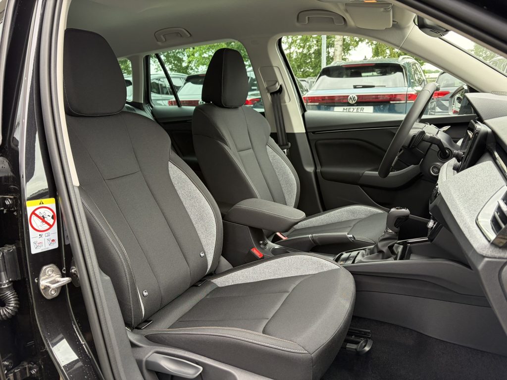 Interior of a right-hand-drive car showing two black fabric front seats, center console, and steering wheel, with parked cars visible outside through the windows.