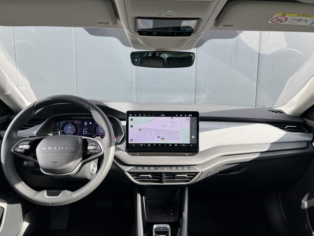 Front interior of a Skoda car with steering wheel, large central touchscreen showing navigation, and rearview mirror visible above the dash.