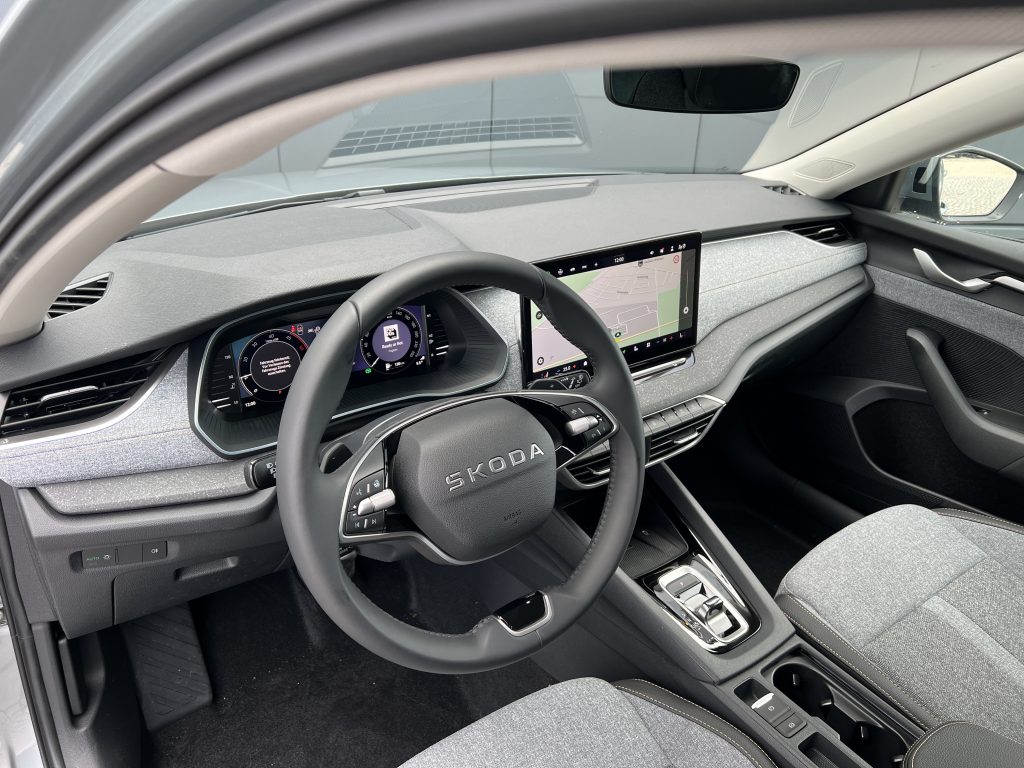 Modern car cockpit with a digital instrument cluster and a central touchscreen displaying a navigation map, viewed from the driver's seat (Skoda branding on the steering wheel).