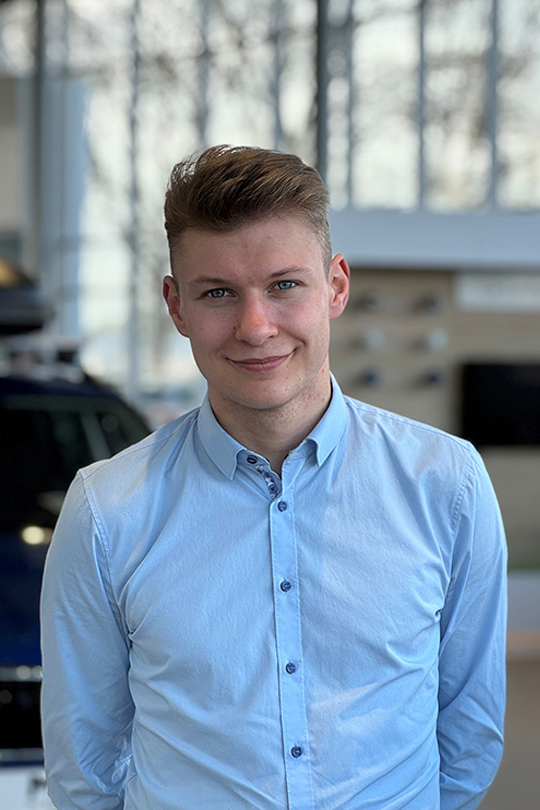 Portrait of a young man in a light blue button-down shirt, smiling, in a showroom with cars in the background.