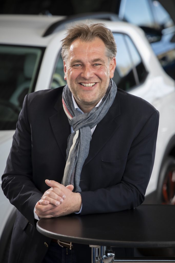 Smiling man in a dark blazer and scarf leaning on a round black table, with a car in the background