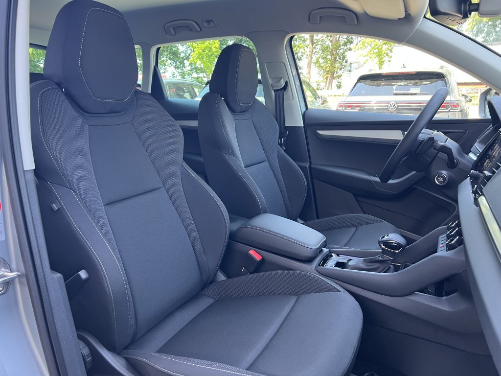 Interior of a car with two front seats, dark fabric upholstery and white contrast stitching, steering wheel and center console visible outside light from windows.