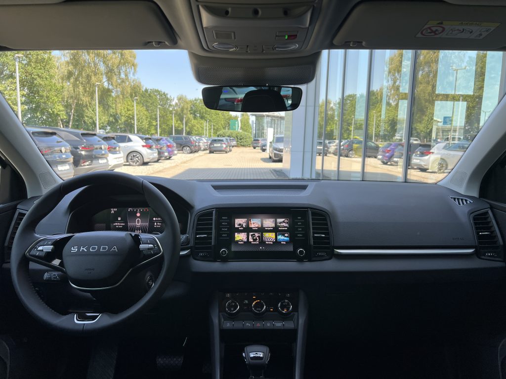 Driver’s perspective inside a Skoda: steering wheel, central touchscreen, and overhead console, with a parking lot seen through the windshield and side window.