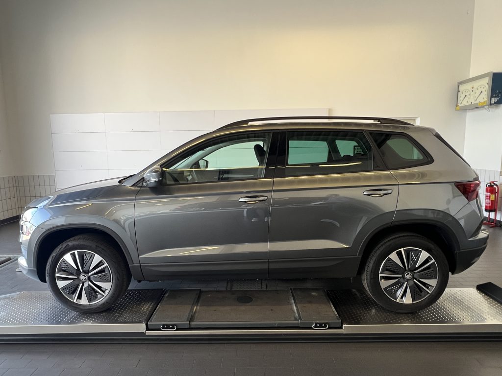 Grey SUV on a car lift in a showroom, side view with roof rails visible against light walls and tiled lower walls.