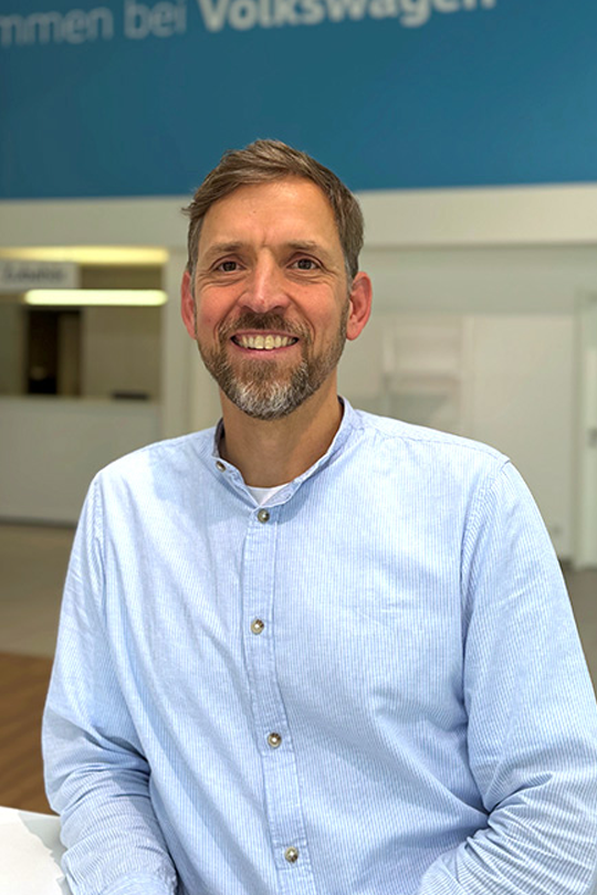 Smiling man with short hair and a beard, wearing a light blue button-down shirt, seated indoors with a blue wall and reception area in the background.