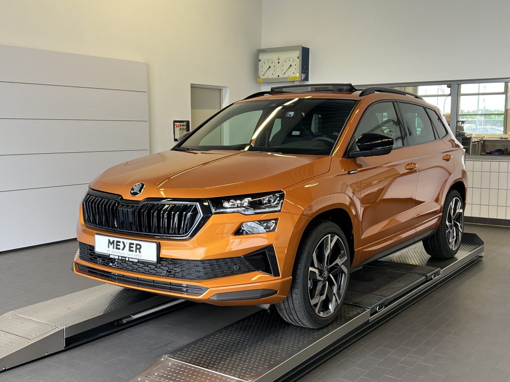 Orange SUV positioned on a metal roller testing platform inside a showroom, front-left view visible