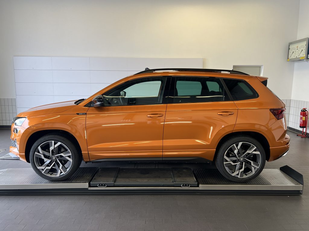 Orange SUV on a raised inspection platform inside a bright showroom or service bay, with roof rails and large alloy wheels visible in the foreground.
