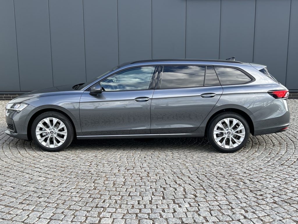 Side view of a silver station wagon parked on a cobblestone street beside a grey panel wall.