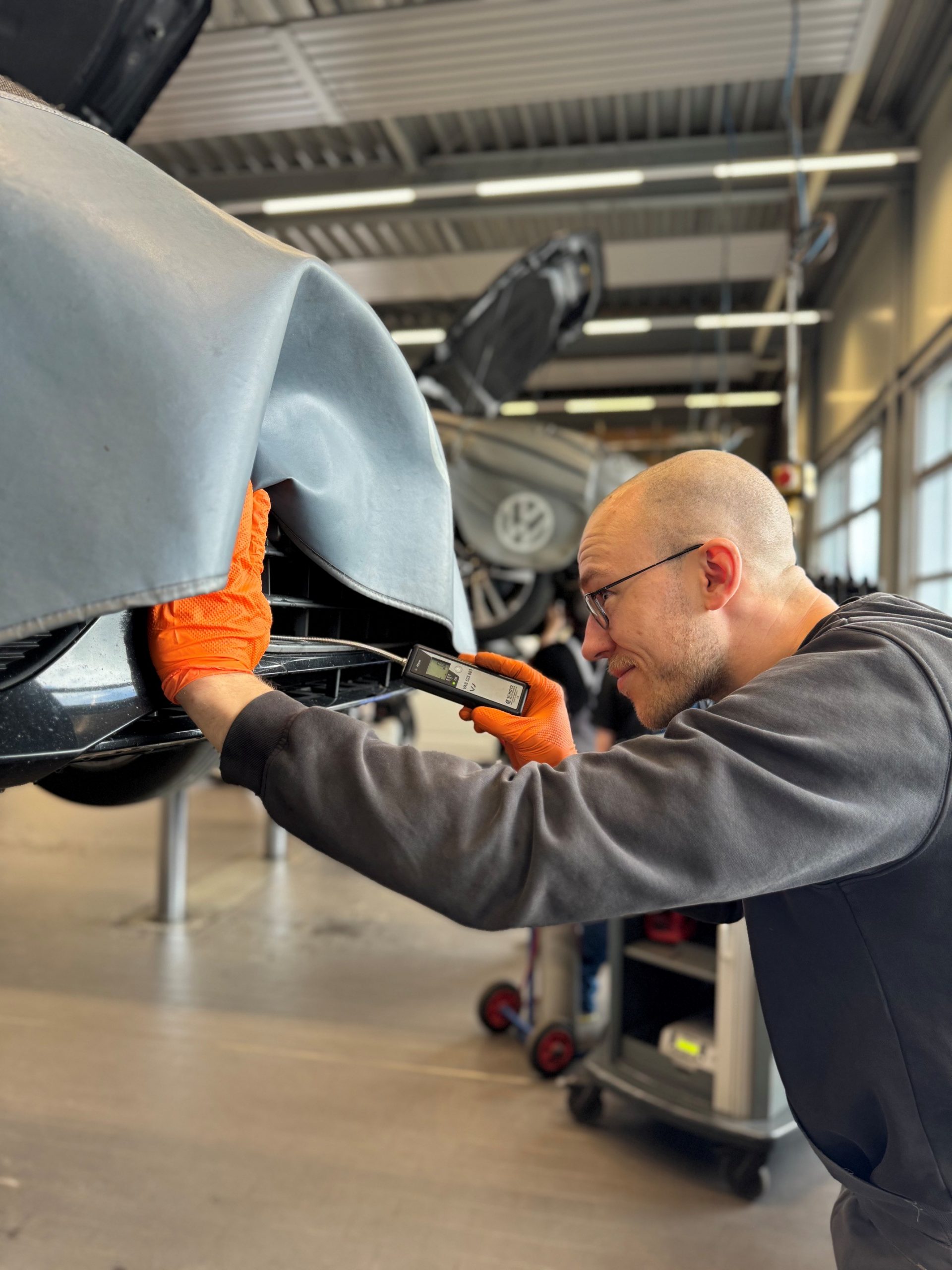 Male mechanic wearing orange gloves uses a handheld diagnostic tool on a car's front bumper in a workshop.