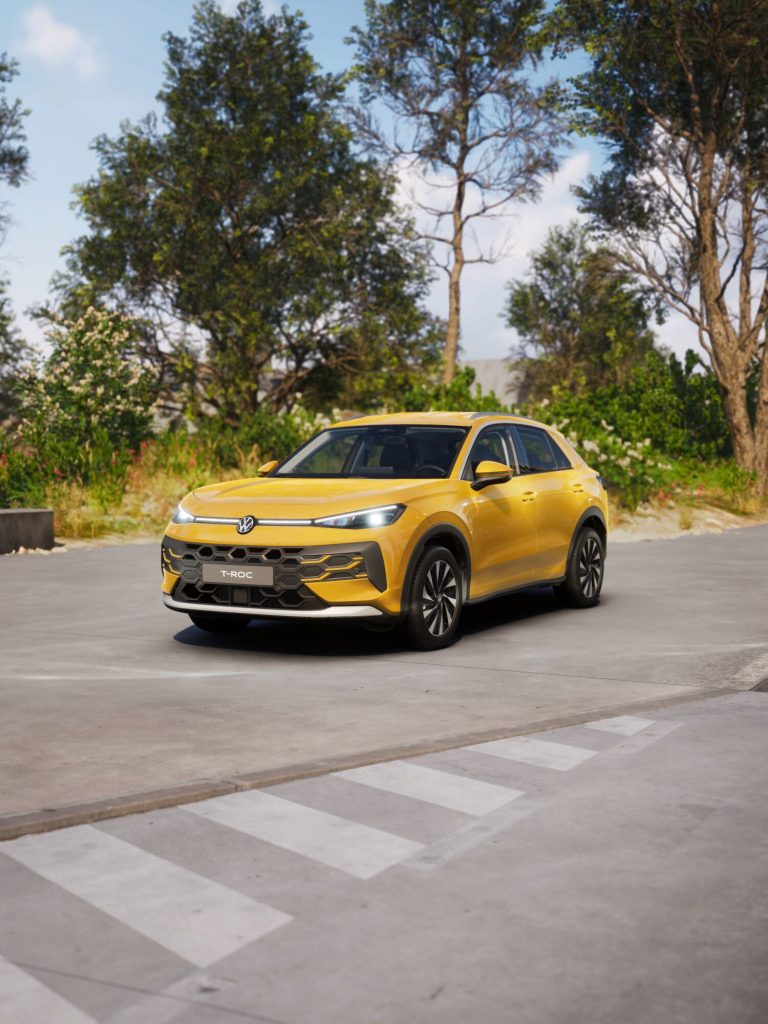 Front three-quarter view of a yellow Volkswagen T-Roc SUV parked on a paved street with a crosswalk in the foreground and trees behind fit.