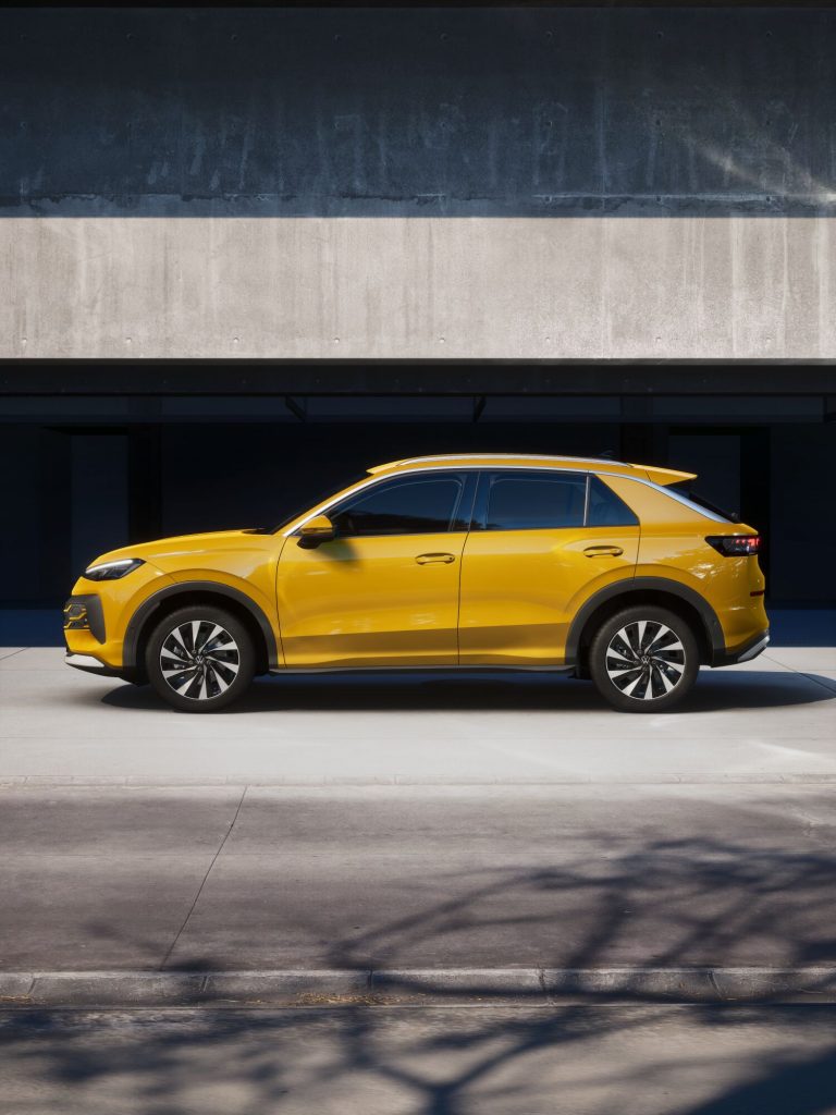 Side view of a bright yellow SUV parked under a dark concrete overhang, with a pale pavement in front.