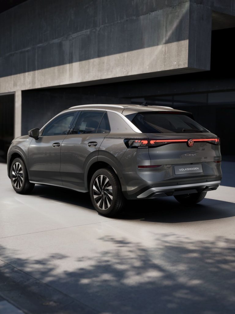 Rear three-quarter view of a gray Volkswagen T-Roc SUV parked beside a modern concrete building, showing the distinctive taillight bar.