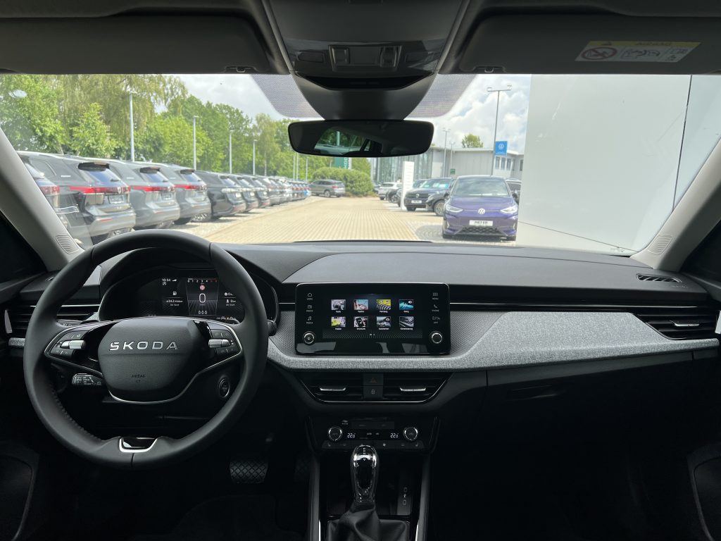 Interior view of a Skoda showing steering wheel, digital instrument cluster, and a large central touchscreen, with a dealership parking lot outside.