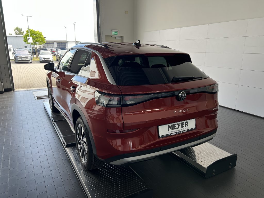 Rear three-quarter view of a burgundy VW T‑Roc SUV on a metal display platform inside a showroom, license plate Meyer Autohaus visible outside.