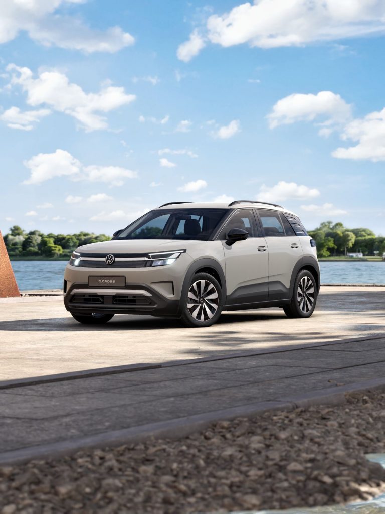 Front three-quarter view of a light gray VW ID. Cross SUV parked by a waterfront with a blue sky and scattered clouds in the background.