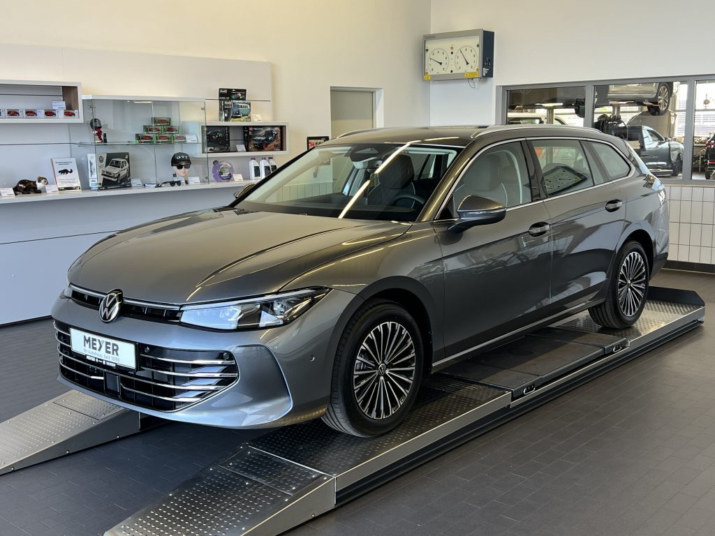 Gray Volkswagen sedan on a display lift inside a car showroom, with product shelves and a wall clock in the background